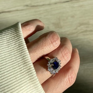 May include: A silver ring featuring a large, round, dark blue gemstone surrounded by smaller, clear, baguette-cut stones. The ring is set on a hand wearing a white sweater.
