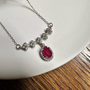 Könnte beinhalten: Eine silberfarbene Halskette mit einer feinen Kette, die mit einer Reihe von vierblättrigen Blumenmotiven verbunden ist, welche mit kleinen klaren Steinen besetzt sind. Ein zentraler tropfenförmiger dunkelroter Edelstein ist von einem Kranz aus kleinen klaren Steinen umgeben.
