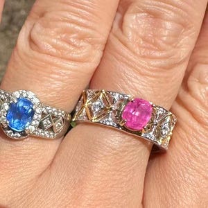 Könnte beinhalten: Zwei Ringe mit oval geschliffenen Edelsteinen. Der linke Ring hat einen blauen ovalen Stein, der von kleinen weißen Steinen umgeben ist. Der rechte Ring hat einen rosa ovalen Stein in einer Ringschiene mit goldenen geometrischen Akzenten.
