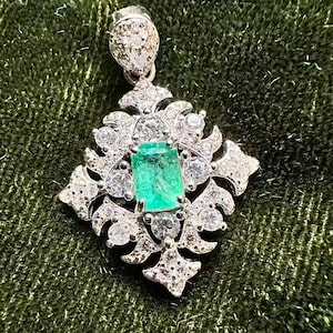 Könnte beinhalten: Ein silberfarbener Anhänger mit einem zentralen, smaragdgeschliffenen grünen Edelstein, umgeben von einem dekorativen Filigranrahmen, der mit zahlreichen kleinen, klaren, rund geschliffenen Steinen besetzt ist. Eine birnenförmige Öse mit klaren Steinen sitzt oben.