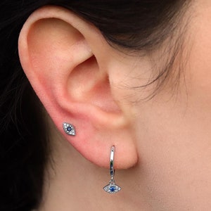 May include: A pair of silver earrings featuring an eye design. One is a small stud earring, and the other is a hoop earring with a dangling charm. Both earrings have a blue center and are surrounded by small, clear stones.