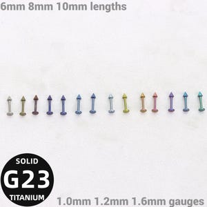 May include: A collection of fourteen metal labret studs with cone-shaped ends in various colours, including silver, gold, blue, purple, and green. Text reads 6mm 8mm 10mm lengths, SOLID G23 TITANIUM, and 1.0mm 1.2mm 1.6mm gauges.