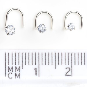 May include: Three silver nose rings with a clear cubic zirconia stone. The rings are different sizes and are shown on a ruler with measurements in millimeters and centimeters.