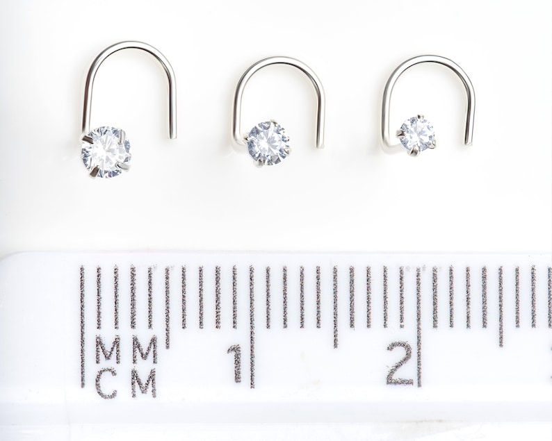 May include: Three silver nose rings with a clear cubic zirconia stone. The rings are different sizes and are shown on a ruler with measurements in millimeters and centimeters.