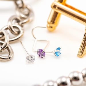 May include: A close-up of three pairs of earrings with different colored gemstones. The earrings are silver and feature a clear, purple, and blue gemstone. The earrings are on a white surface.