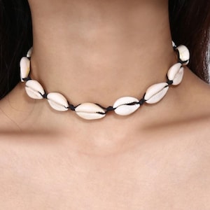 May include: A white seashell choker necklace with black string. The necklace is made of small, oval-shaped seashells strung together on a black cord.