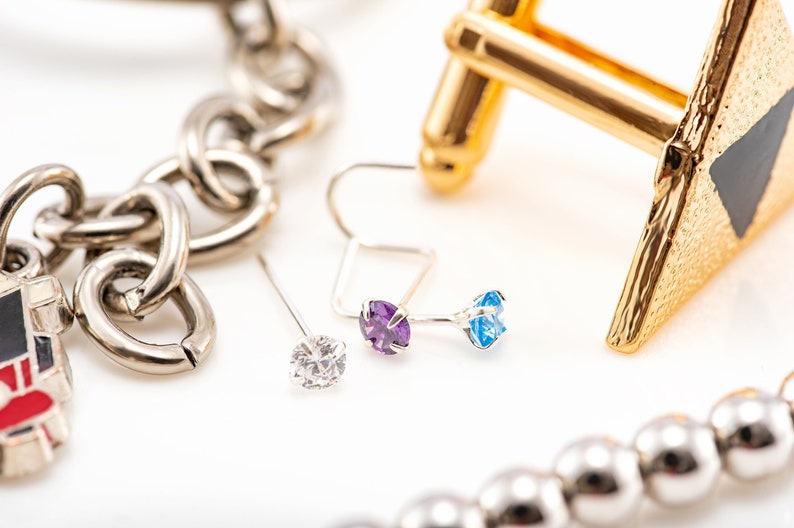 May include: A close-up of three silver earrings with different colored gemstones. The earrings are on a white surface. The earrings have a clear, purple, and blue gemstone.