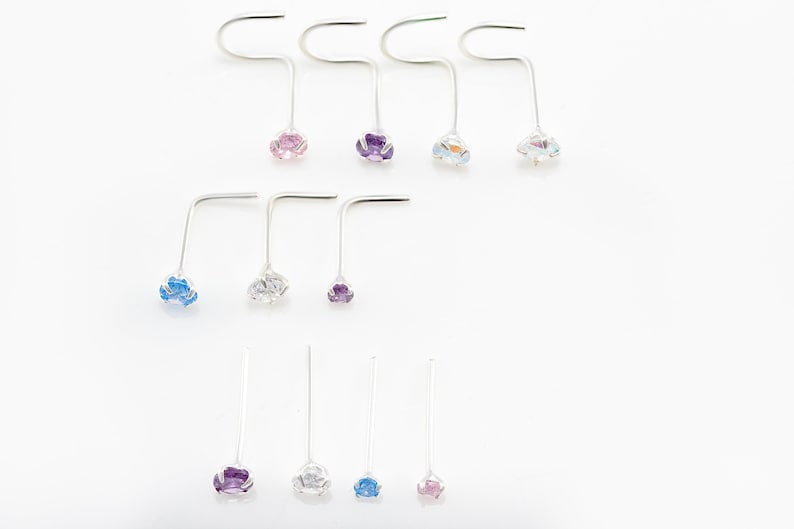 May include: Assortment of nose rings with different colored gemstones. The rings are made of silver and have a curved shape. The gemstones are blue, purple, and pink.