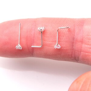 May include: Three silver nose rings with clear rhinestones. The rings are on a finger.