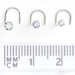 May include: Three silver nose rings with clear cubic zirconia stones. The rings are shown next to a ruler, with the smallest ring measuring 5 mm, the middle ring measuring 7 mm, and the largest ring measuring 9 mm.