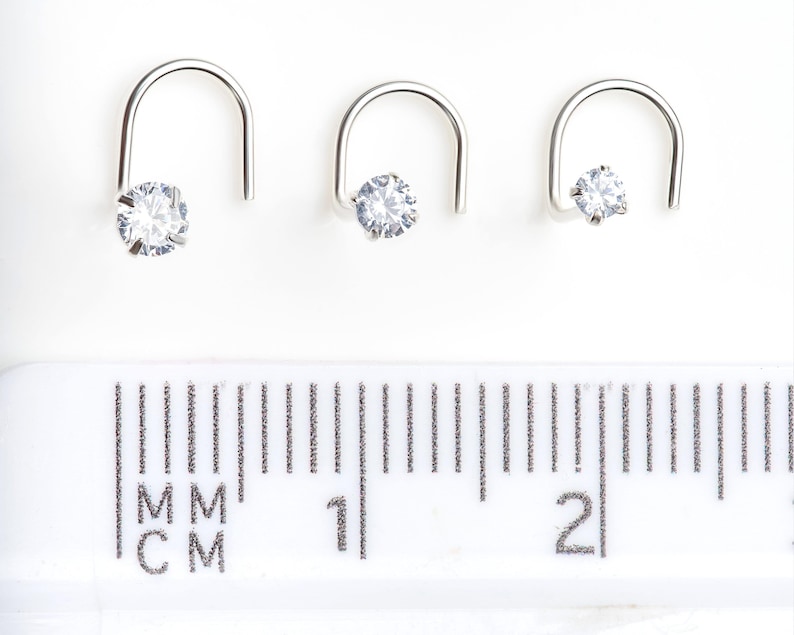 May include: Three silver nose rings with clear cubic zirconia stones. The rings are shown next to a ruler, with the smallest ring measuring 5 mm, the middle ring measuring 7 mm, and the largest ring measuring 9 mm.