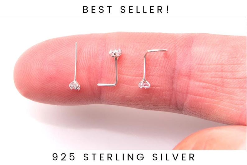 May include: Three silver nose rings with a small clear gemstone on each. The rings are displayed on a finger.
