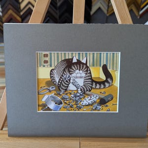 May include: A framed print featuring a cartoon cat surrounded by watch parts. The cat, with gray and brown stripes, appears to be working on a watch. The print is matted in gray and displayed on a wooden easel.