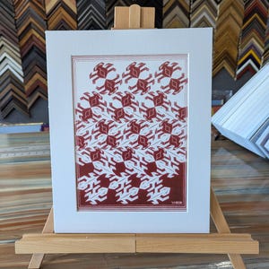 May include: A framed print featuring a repeating pattern of red and white geometric shapes. The artwork is displayed on a small wooden easel, with a white mat and a red border. The background includes various picture frames.