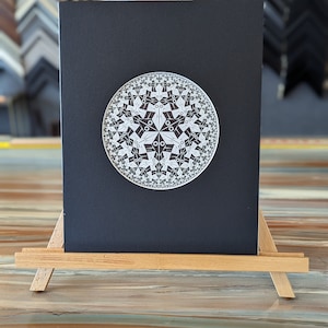 May include: A black and white geometric design printed on a black card, displayed on a wooden easel.