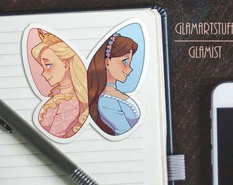 May include: A sticker featuring two cartoon women with butterfly wings. The woman on the left has blonde hair and is wearing a pink and white dress. The woman on the right has brown hair and is wearing a blue dress. The sticker is on a notebook with the text "Glamartstuff" and "Glamist" written on the cover.