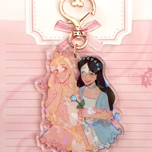 May include: A clear acrylic keychain featuring two cartoon women in princess dresses. The woman on the left has blonde hair and is wearing a pink dress with a blue rose. The woman on the right has dark hair and is wearing a blue dress with a floral crown. Both women are smiling and holding hands. The keychain has a gold heart-shaped clasp.