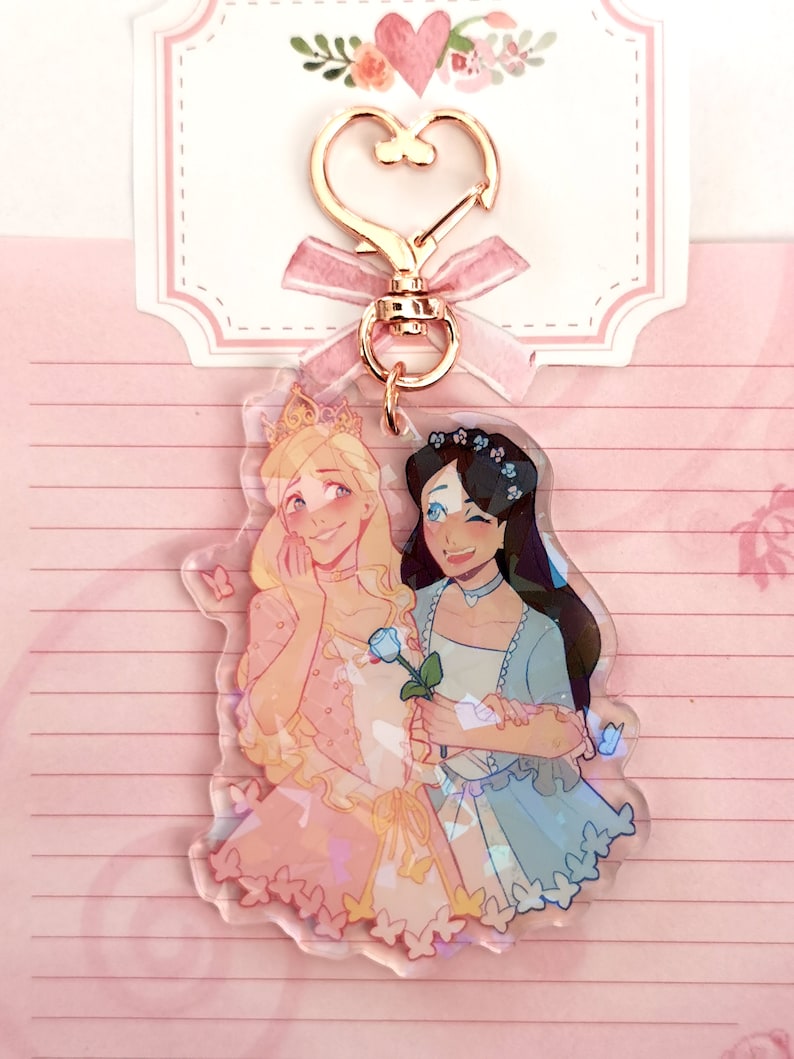 May include: A clear acrylic keychain featuring two cartoon women in princess dresses. The woman on the left has blonde hair and is wearing a pink dress with a blue rose. The woman on the right has dark hair and is wearing a blue dress with a floral crown. Both women are smiling and holding hands. The keychain has a gold heart-shaped clasp.