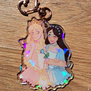 May include: A holographic charm featuring two women in princess dresses, one in pink and one in blue, holding a blue rose. The charm has a gold heart-shaped keychain.