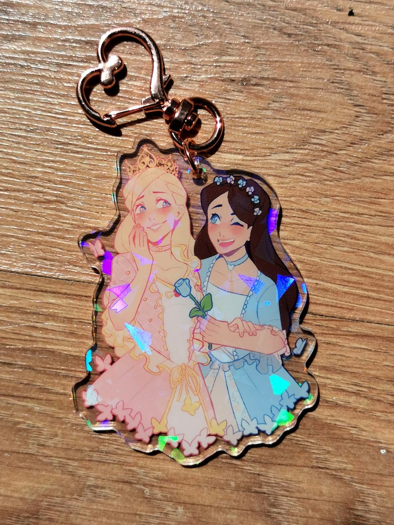 May include: A holographic charm featuring two women in princess dresses, one in pink and one in blue, holding a blue rose. The charm has a gold heart-shaped keychain.