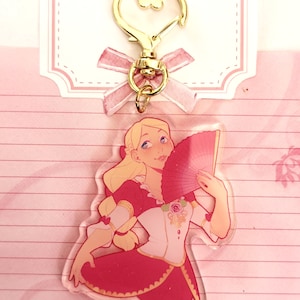 May include: A pink and gold charm featuring a cartoon woman with blonde hair holding a red fan. The charm is attached to a gold heart-shaped keychain.