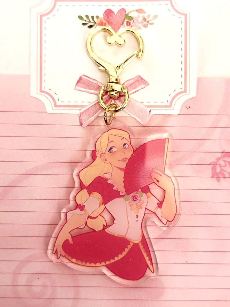 May include: A pink and gold charm featuring a cartoon woman with blonde hair holding a red fan. The charm is attached to a gold heart-shaped keychain.