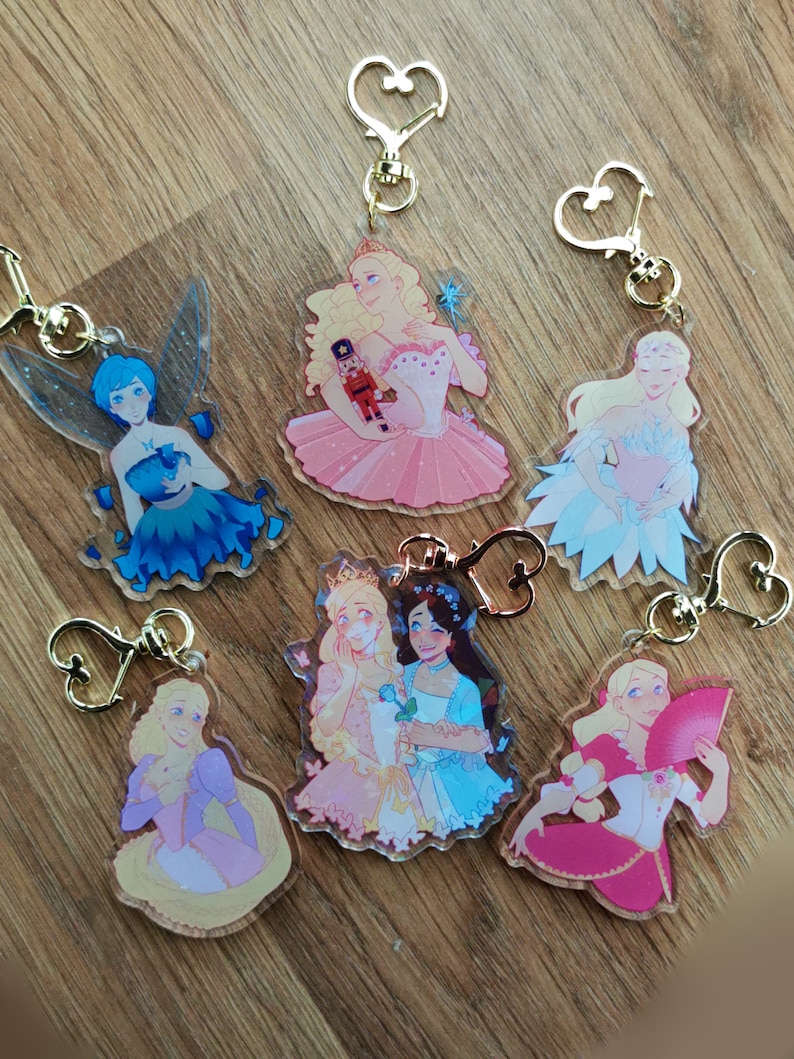 May include: Six clear acrylic keychains with different illustrations of women in various poses and outfits. The keychains feature gold-colored metal heart-shaped rings.
