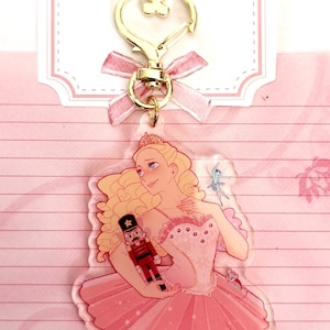 May include: A clear acrylic keychain featuring a ballerina in a pink tutu holding a nutcracker doll. The ballerina has blonde hair and is wearing a tiara. The keychain has a gold heart-shaped clasp.