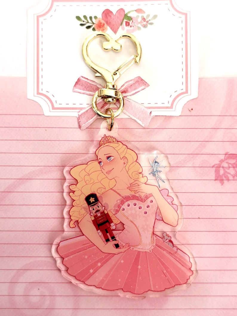 May include: A clear acrylic keychain featuring a ballerina in a pink tutu holding a nutcracker doll. The ballerina has blonde hair and is wearing a tiara. The keychain has a gold heart-shaped clasp.
