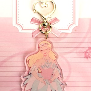 May include: A clear acrylic keychain featuring a cartoon illustration of a blonde woman in a pink and blue dress. The keychain has a gold heart-shaped clasp and a pink ribbon.