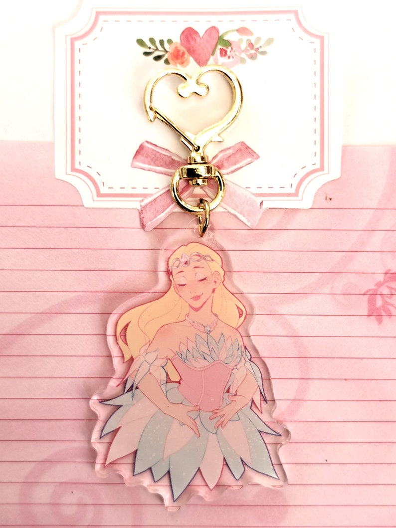 May include: A clear acrylic keychain featuring a cartoon illustration of a blonde woman in a pink and blue dress. The keychain has a gold heart-shaped clasp and a pink ribbon.