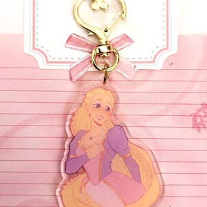 May include: A clear acrylic keychain featuring a cartoon illustration of a blonde woman with long hair, wearing a pink and purple dress. The woman is smiling and holding a small red heart in her hand.
