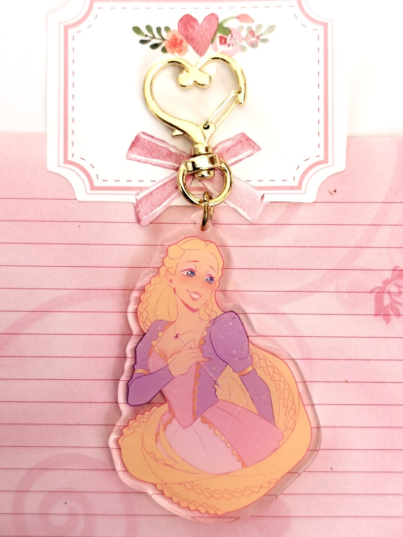 May include: A clear acrylic keychain featuring a cartoon illustration of a blonde woman with long hair, wearing a pink and purple dress. The woman is smiling and holding a small red heart in her hand.