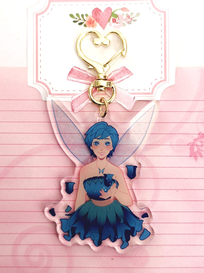May include: A blue fairy with wings and a blue flower dress holding a blue flower. The fairy is a keychain with a gold heart-shaped clasp.