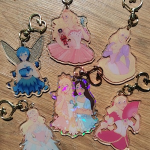May include: Seven colorful keychains featuring different characters from the Nutcracker ballet. Each keychain is made of clear acrylic with a holographic finish and features a different character from the ballet, including the Sugar Plum Fairy, the Nutcracker, and Clara. The keychains are attached to gold heart-shaped rings.