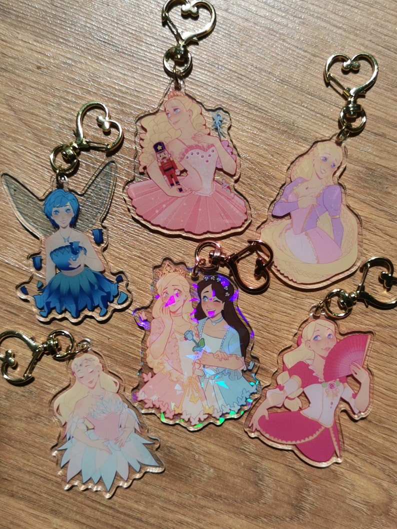 May include: Seven colorful keychains featuring different characters from the Nutcracker ballet. Each keychain is made of clear acrylic with a holographic finish and features a different character from the ballet, including the Sugar Plum Fairy, the Nutcracker, and Clara. The keychains are attached to gold heart-shaped rings.