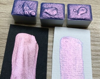 May include: Three small square containers of iridescent metallic paint. The paint is a light purple color with a shimmery finish. The paint is shown on a black surface and a white surface.
