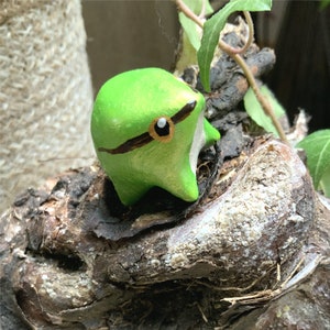 Könnte beinhalten: Eine kleine grüne Froschfigur mit schwarzen Augen und einem schwarzen Streifen über den Augen, die auf einem Stück verwittertem Holz sitzt.