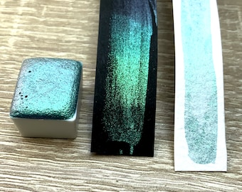 May include: A close-up of a teal green metallic watercolor paint in a small square pan, a swatch of the paint on a black paper strip, and a swatch of the paint on a white paper strip.