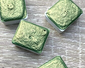 May include: Four square watercolor paint pans with a metallic green pigment. The pans are white with a slightly raised edge around the paint.
