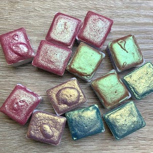 Könnte beinhalten: Eine Sammlung quadratischer Aquarellfarben in verschiedenen schillernden Farben, darunter Rosa, Grün und Blau. Jede Farbe befindet sich in einem weißen Kunststoffhalter. Die Farben haben ein schimmerndes, metallisches Finish, was auf eine künstlerische Verwendung hindeutet.