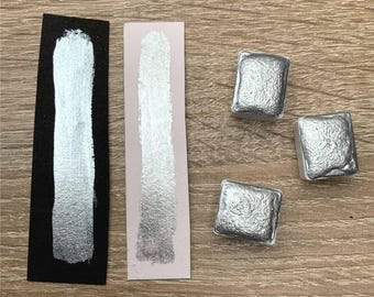 May include: Three silver watercolor paint pans, two on a white background and one on a black background. The paint pans are square and have a metallic sheen.