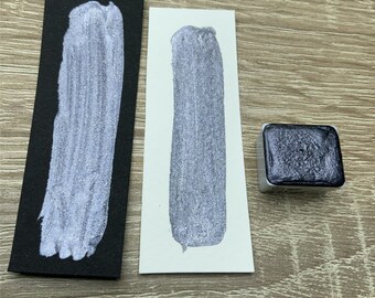 May include: A silver watercolor paint swatch on black paper and white paper, next to a silver paint pan. The paint has a metallic sheen. The paint is on a wooden surface.