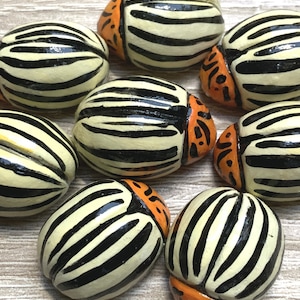 Könnte beinhalten: Neun Keramikkäfer, die mit schwarzen und weißen Streifen und orangefarbenen Akzenten bemalt sind. Die Käfer sind in einer Gruppe auf einer hellbraunen Holzoberfläche angeordnet.
