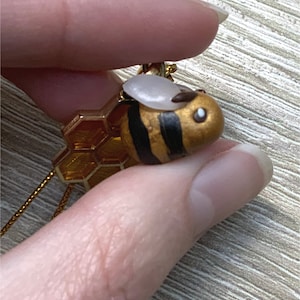 Könnte beinhalten: Eine goldene Bienen-Anhängerkette mit einem Honigwaben-Anhänger. Die Biene ist mit schwarzen Streifen bemalt und hat ein weißes Gesicht mit schwarzen Augen.
