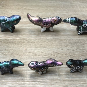 Könnte beinhalten: Sechs kleine, handbemalte Figuren von Eidechsen mit schillerndem grünen, violetten und schwarzen Lack. Die Eidechsen haben weiße Akzente und sind in verschiedenen Positionen aufgestellt.