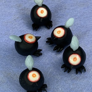 Könnte beinhalten: Fünf schwarze und weiße Tonfiguren mit einem roten Auge und einem kleinen blauen Flügel. Die Figuren stehen auf einer blauen Oberfläche.