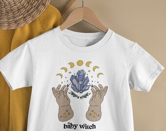 May include: White t-shirt with a graphic featuring a blue crystal, crescent moons, and hands. The text "baby witch" is printed below the graphic. The shirt is hanging on a wooden hanger.