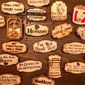 May include: A collection of laser-cut wooden signs with various humorous phrases and quotes. The signs feature text like "I'm not short, I'm a Hobbit" and "I could out pizza the hut." The signs are arranged on a wooden surface.
