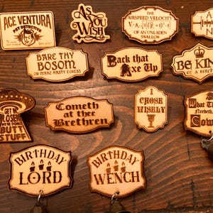 May include: A collection of wooden plaques with various engraved phrases and images. The plaques feature text like "Ace Ventura Pet Detective", "As You Wish", and "Birthday Lord". Some plaques have small bells attached. The overall aesthetic is rustic and humorous.